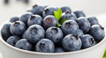 Primer plano de un tazón lleno de arándanos azules redondos con una pequeña hoja verde, destacando su superficie cerosa y la forma de su corona.
