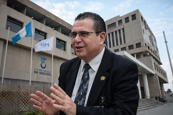 Hombre con traje y gafas gesticula frente a un edificio. Detrás, una bandera de Guatemala, otra con el logo "CIM" y texto "MP Certificación ISO 9001-2015".