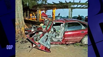 Un automóvil de color rojo destruido junto a un árbol, con una estructura con columnas y policías en el fondo