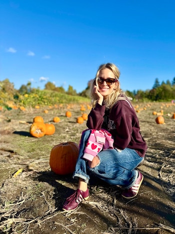 Una mujer con gafas de sol se agacha en un campo de tierra junto a varias calabazas anaranjadas bajo un cielo despejado