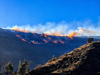 Fuego y columnas de humo cubren la cresta de una ladera montañosa frente a un terreno seco con vegetación rala y cielo azul