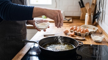 Una persona añade harina a una sartén sobre una placa de cocina, junto a un tazón con harina y un cartón de huevos.