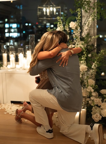Una mujer rubia y un hombre arrodillados se abrazan en una habitación decorada con arreglos de flores blancas, velas encendidas y pétalos