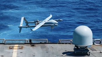 Un dron gris de alas fijas vuela bajo sobre las aguas azules del océano, cerca de la cubierta de un barco gris con una cúpula de radar blanca
