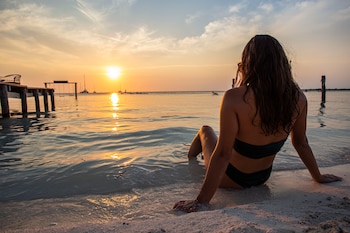 Mujer de espaldas con cabello oscuro y traje de baño sentada en la arena con agua. El sol se refleja en el mar, se ven un muelle y botes