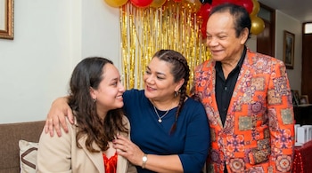 Tres personas sonrientes en una sala decorada con globos. Melcochita, un hombre mayor, viste un saco naranja. Una mujer de mediana edad y una joven a su lado.