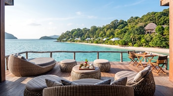 Muebles de mimbre sobre una terraza de madera con vista al mar turquesa, una playa de arena blanca y árboles.