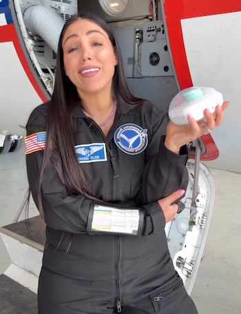 Una mujer con traje de vuelo negro sostiene un dispositivo blanco frente a la compuerta abierta de una aeronave