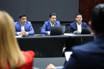 Tres hombres vestidos con trajes azules sentados a una mesa oscura en una sala de conferencias. Hay micrófonos y ordenadores portátiles