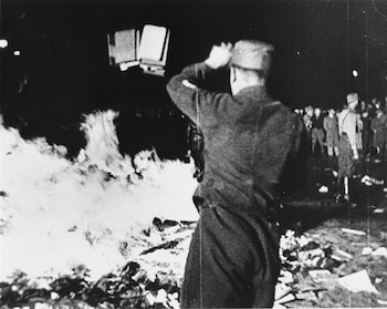 Hombre de uniforme lanza libros a una gran hoguera por la noche. Las llamas iluminan los escombros y a una multitud de personas en el fondo oscuro