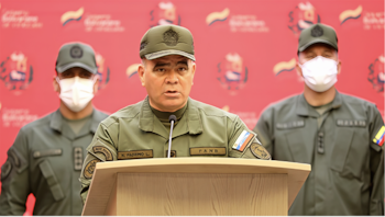Un oficial militar habla tras un podio mientras dos uniformados con mascarillas permanecen de pie detrás, sobre un fondo rojo institucional