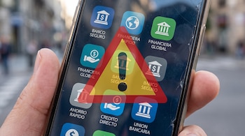 Mano sostiene celular de pantalla quebrada, que exhibe íconos de aplicaciones bancarias y un símbolo de advertencia triangular con signo de exclamación.