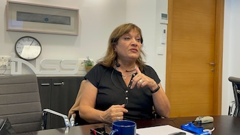 Mujer con micrófono de diadema sentada en un escritorio, señalando con la mano. Detrás hay un reloj, un cartel INSS y una puerta de madera