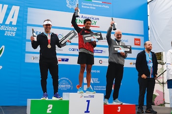 Tres corredores masculinos posan en un podio con los números 1, 2 y 3, levantando botellas y mostrando medallas, con un fondo azul de patrocinadores y "MaraTANA 2026"