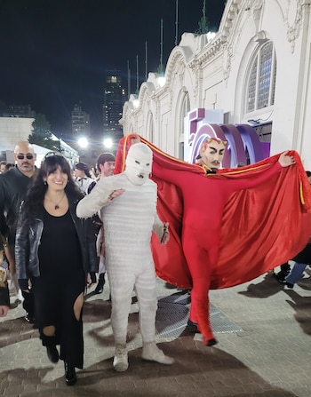 Una mujer, un hombre disfrazado de momia y otro de diablo con capa roja caminan de noche por un sendero adoquinado junto a un edificio blanco