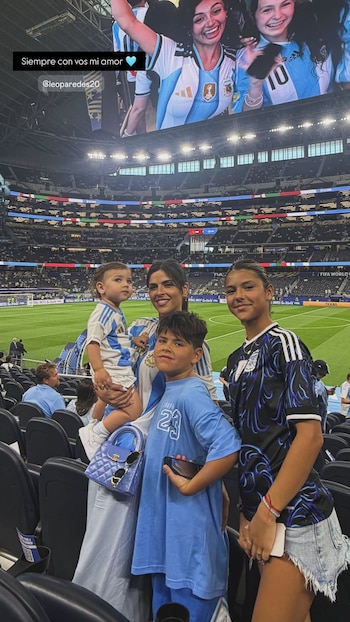 Mujer con bebé en brazos, dos niños en gradas de estadio de fútbol. Visten camisetas de Argentina. Pantalla gigante con dos mujeres sonrientes en el fondo