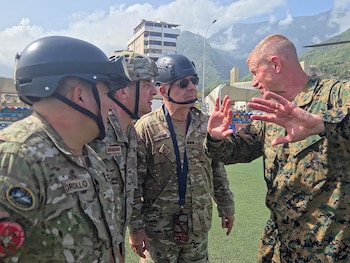 Cuatro hombres con uniformes militares y cascos están de pie al aire libre, un hombre habla con gestos de manos; montañas y edificios al fondo