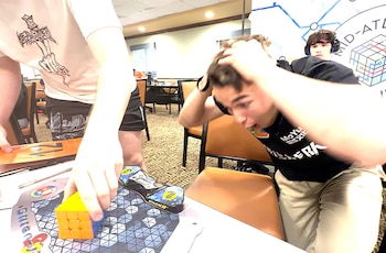 Hombre joven con manos en la cabeza reacciona. Otro hombre coloca un cubo Rubik sobre alfombrilla y temporizador digital. Al fondo, otro participante con auriculares