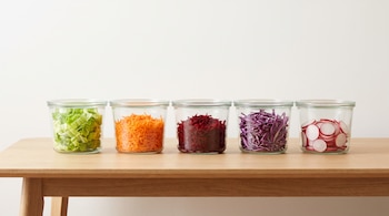Mesa de madera con cinco recipientes herméticos de vidrio alineados, cada uno contiene lechuga, zanahoria, remolacha, repollo colorado y rabanitos cortados.
