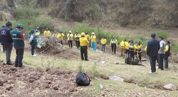 Un grupo de personas con camisetas amarillas y palas escucha a instructores con chalecos en una zona rural con ramas secas