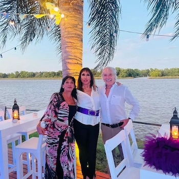 Tres personas posan junto a una palmera y mesas blancas en una terraza situada a orillas del agua