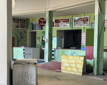 Interior de un restaurante de pizza con paredes verdes y temática de selva. Se ven mostradores, menús, un congelador sucio y escombros. Una ventana está al fondo