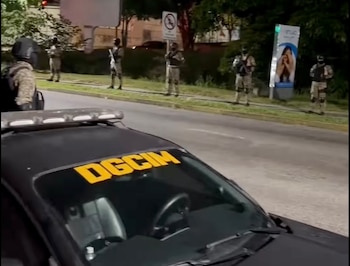 Coche negro con "DGCIM" en el parabrisas y barra de luces en el techo, estacionado de noche frente a militares en uniforme camuflado junto a una calle