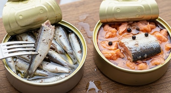 Dos latas de conserva abiertas con sardinas y salmón sobre una tabla de madera. Un tenedor sostiene una sardina.