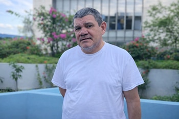 Retrato de un hombre de mediana edad, con cabello y barba grises, vistiendo una camiseta blanca. El fondo incluye plantas, flores y un edificio moderno.