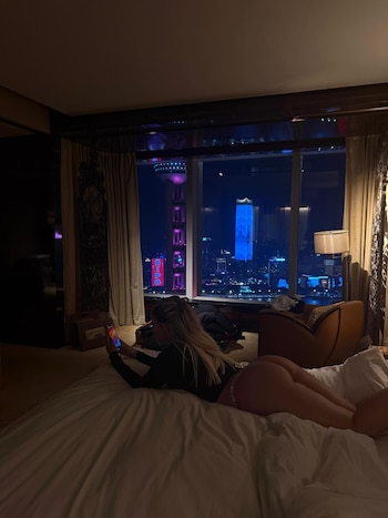 Una mujer recostada en una cama sostiene un teléfono móvil frente a una gran ventana que da a una ciudad iluminada de noche