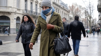 Una mujer con abrigo verde, gorro gris, bufanda azul y beige, y guantes marrones camina por una calle urbana con edificios antiguos al fondo.