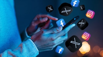 Una mano toca la pantalla de un teléfono móvil, rodeada de iconos flotantes de X, Facebook e Instagram, con un fondo oscuro y luces desenfocadas.