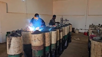 Dos hombres trabajan en un taller rodeados de filas de cilindros metálicos, uno de ellos soldando con gafas de protección