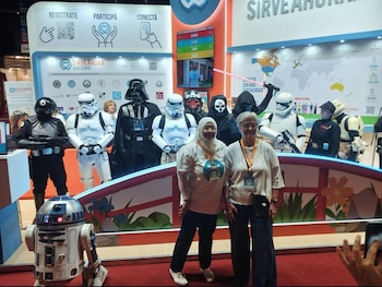 Dos mujeres sonríen frente a personajes de Star Wars, incluyendo Darth Vader, Stormtroopers, un soldado imperial, y R2-D2, en un stand de convención