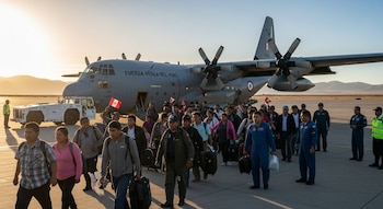 Personas con equipaje y banderas peruanas caminan por una pista de aeropuerto al atardecer, con un avión militar gris grande de la Fuerza Aérea del Perú detrás y montañas.