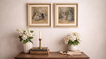 Dos cuadros con grabados de escenas de figuras y marcos dorados cuelgan en una pared. Sobre una mesa de madera se ven jarrones con flores, libros y un candelabro.