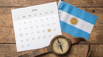 Composición plana: calendario de junio de 2026 con los días 15 y 20 marcados, bandera argentina y brújula vintage sobre escritorio de madera.