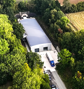 Vista aérea de un edificio industrial, un túnel de piedra, un camión azul, coches estacionados, densa vegetación y un campo de cultivo