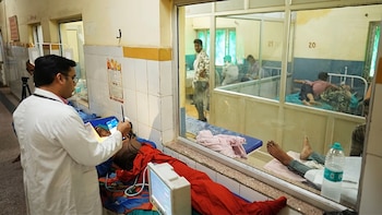 Un médico con bata blanca atiende a un paciente recostado. Se observan equipos médicos, un monitor, y una ventana que muestra otros pacientes en una sala