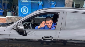 Un hombre dentro de un automóvil negro saluda con la mano mientras sonríe frente a una tienda con camisetas del Cruz Azul.