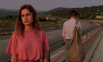 Primer plano de una mujer joven con cabello rizado y camiseta a rayas rojas y blancas, junto a un hombre de espaldas con una bolsa, en el arcén de una carretera