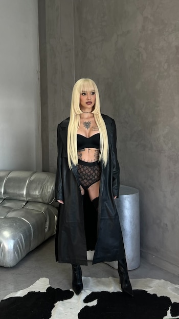 La Joaqui, de pie, con cabello rubio largo, posa con un sostén negro, lencería de encaje, un abrigo largo de cuero negro y botas altas sobre una alfombra de piel de vaca