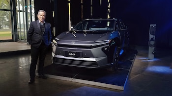 Hombre de traje sonriente junto a un SUV eléctrico gris oscuro en una plataforma. Estación de carga a la derecha y ventanales al fondo