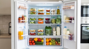 Interior de un refrigerador abierto repleto de frutas, verduras, huevos, botellas de leche y recipientes de vidrio con comida.