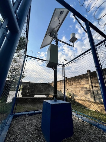 Panel solar, caja eléctrica, poste, sensor, base de concreto azul, valla de malla metálica, alambre de púas, suelo de grava, muro, cielo azul con nubes