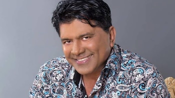 Hombre de piel oscura y cabello oscuro rizado sonríe. Viste una camisa estampada con patrones de colores azules, blancos y negros. El fondo es gris liso