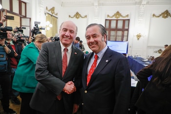 Marinos Kartikkis y Jorge Quiroz, vestidos de traje, se dan la mano y sonríen a la cámara en una sala grande con decoraciones doradas y medios de comunicación