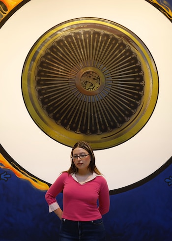 Mujer joven con camiseta rosa y gafas delante de un mural circular. El mural tiene un centro dorado con un rostro y rayos, sobre un fondo azul con llamas