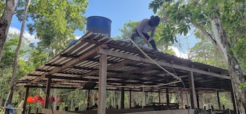 Un hombre vestido de gris sobre un tejado de chapa ondulada de una estructura de madera, con un tanque azul y árboles verdes de fondo
