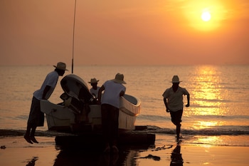 Cuatro hombres con sombreros junto a una pequeña embarcación con motor en la orilla del mar. Uno corre en el agua; dos, cerca del bote. Sol bajo en el horizonte